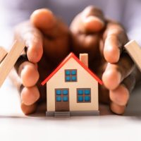 homeowners insurance concept - image of hands around a model house protecting it from falling dominoes