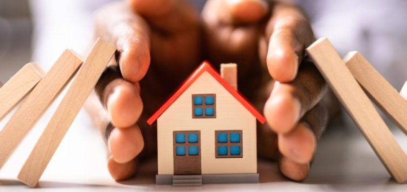 homeowners insurance concept - image of hands around a model house protecting it from falling dominoes