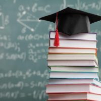 stack of college textbooks with a graduation cap on top in front of a chalkboard full of equations
