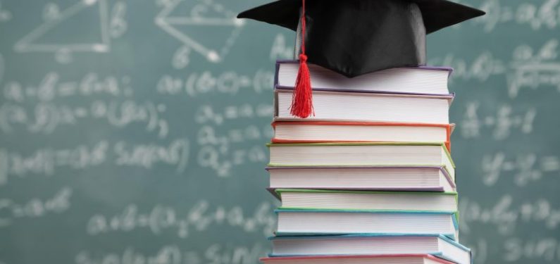 stack of college textbooks with a graduation cap on top in front of a chalkboard full of equations